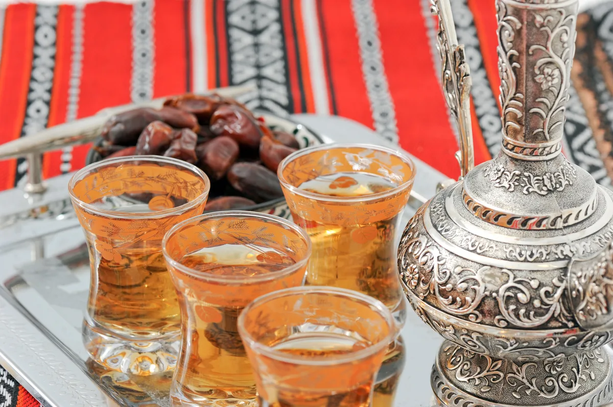 A brass dallah coffee pot pouring cardamom coffee into small cups